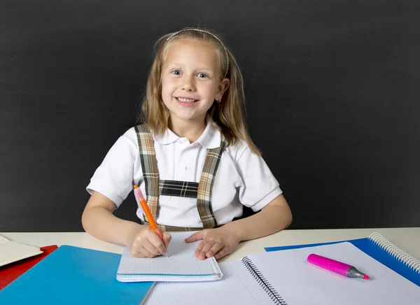 Beautiful happy blond junior schoolgirl smiling while doing school homework writing on notepad with pen
