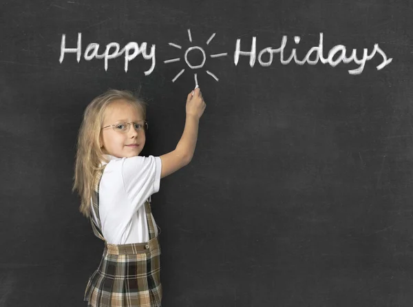 Sweet junior schoolgirl with blonde hair standing happy and smiling writing with chalk happy holidays