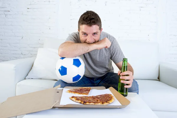 Young man alone holding ball and beer bottle watching football game on television at home sofa couch