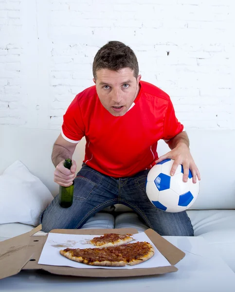 Young man alone holding ball and beer bottle watching football game on television at home sofa couch