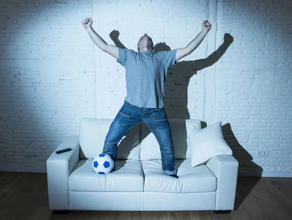 Fanatic and crazy football fan watching television soccer match with ball jumping on sofa celebrating goal
