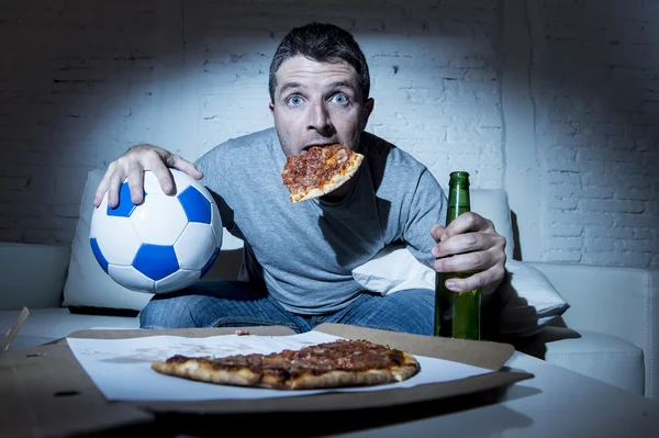 Football fan man watching soccer game on tv at home sofa couch with soccer ball and pizza in his mouth