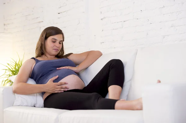 Young beautiful happy 8 or 9 months pregnant woman at home living room couch holding big belly
