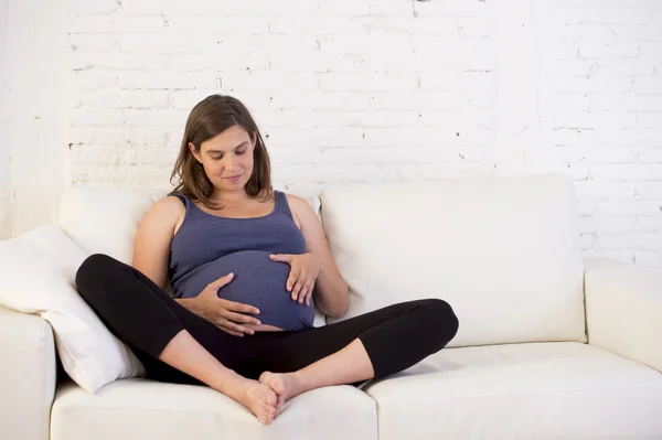 Young beautiful happy 8 or 9 months pregnant woman at home living room couch holding big belly