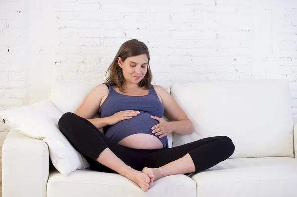 Young beautiful happy 8 or 9 months pregnant woman at home living room couch holding big belly