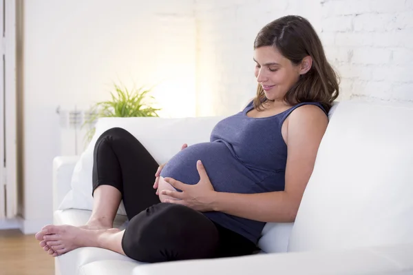 Young beautiful happy 8 or 9 months pregnant woman at home living room couch holding big belly
