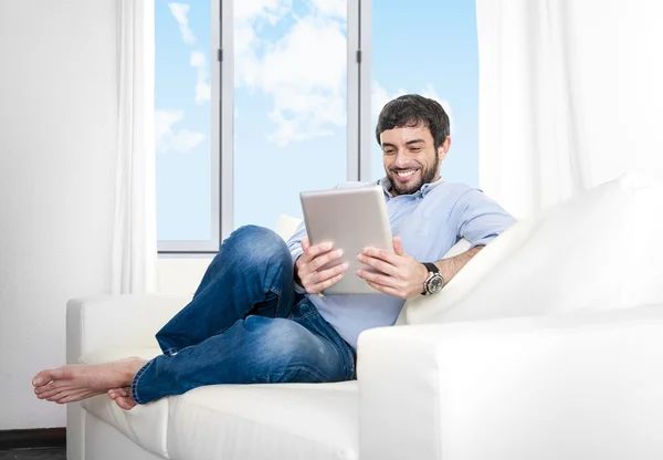 Young attractive Hispanic man at home sitting on white couch using digital tablet