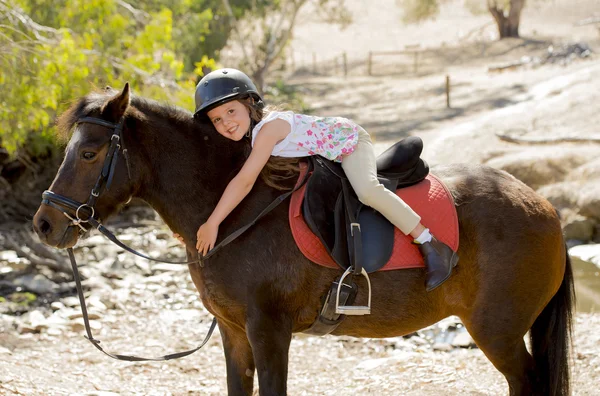Sweet young girl hugging pony horse smiling happy wearing safety jockey helmet in summer holiday