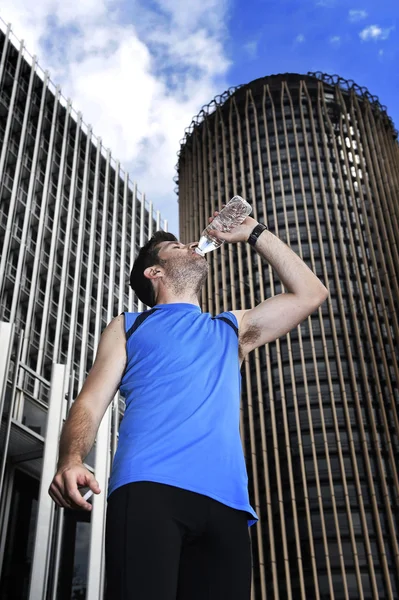 Young sport man drinking water bottle after running training session in business district