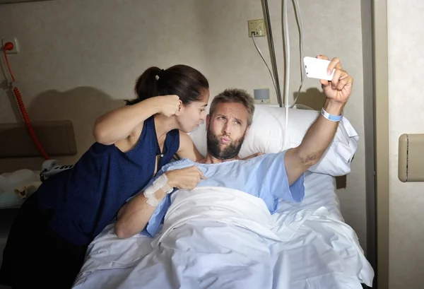 Young couple taking selfie photo at hospital room with man lying in clinic bed