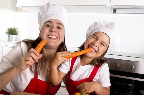 Happy mother and little daughter in apron and cook hat eating carrots together having fun at home kitchen