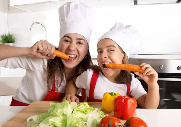 Happy mother and little daughter in apron and cook hat eating carrots together having fun at home kitchen