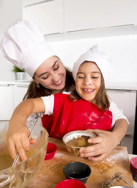 Mother baking with little daughter in apron and cook hat filling mold muffins with chocolate dough