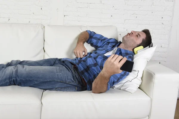 20s or 30s man having fun lying on couch listening to music on mobile phone with headphones playing air guitar