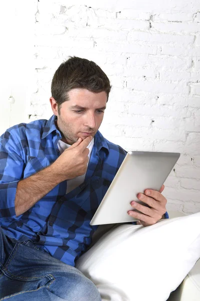 Young attractive 30s man using digital tablet pad lying on couch at home networking looking relaxed