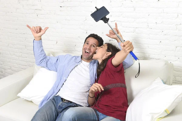 Attractive couple taking selfie photo or shooting self video with mobile phone and stick sitting at home couch smiling happy