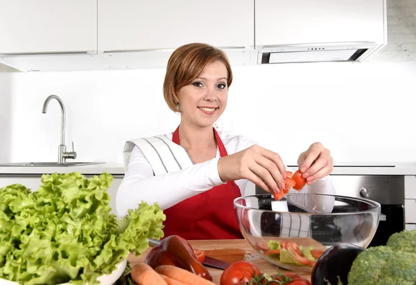 Young beautiful woman in red apron at home kitchen preparing vegetable salad bowl smiling happy