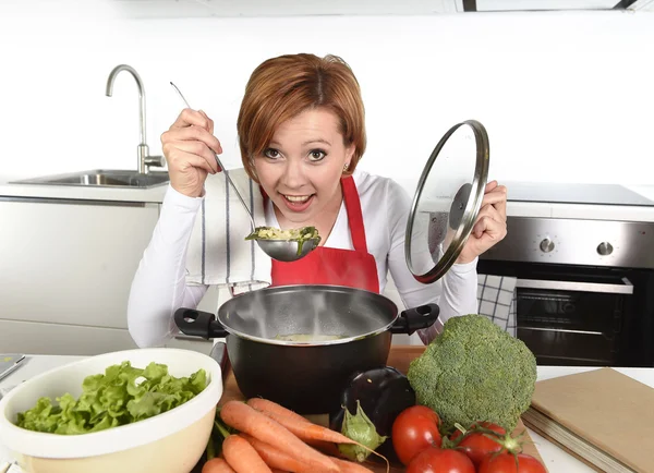 Happy home cook woman in red apron at domestic kitchen holding saucepan with soup tasting delicious vegetable stew