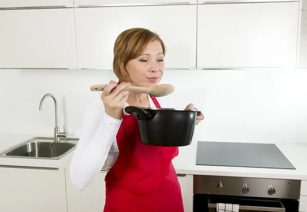 Beautiful home cook woman at kitchen holding spoon and cooking pot tasting delicious soup