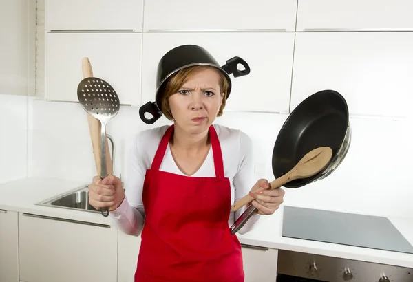 Young attractive home cook woman in red apron at  kitchen holding pan and household with pot on her head in stress