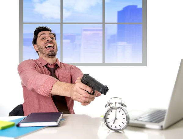 Businessman in stress at office computer pointing hand gun to alarm clock project deadline expiring