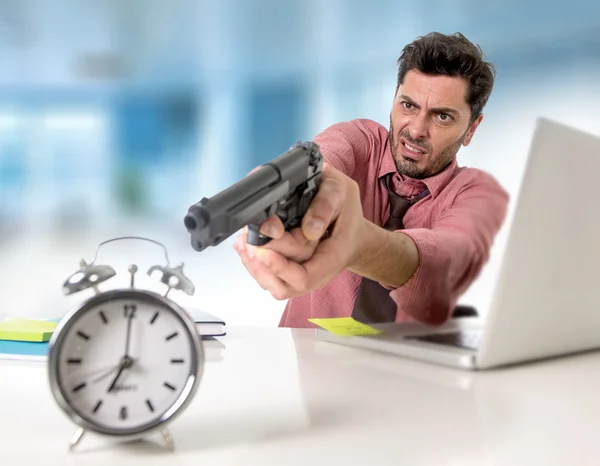Businessman in stress at office computer pointing hand gun to alarm clock project deadline expiring