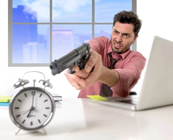 Businessman in stress at office computer pointing hand gun to alarm clock project deadline expiring