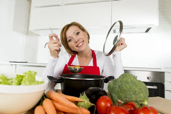 Young happy home cook woman in red apron at domestic kitchen holding saucepan tasting hot soup