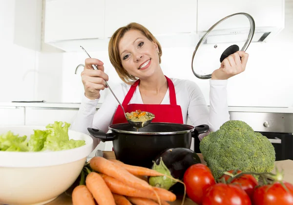 Young happy home cook woman in red apron at domestic kitchen holding saucepan tasting hot soup