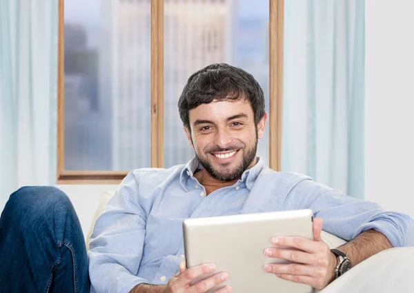 Young attractive Hispanic man at home sitting on white couch using digital tablet