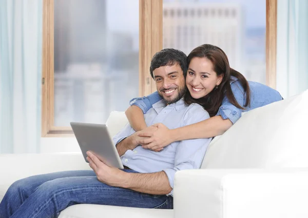 Beautiful latin couple in love lying together on living room sofa couch enjoying using digital tablet