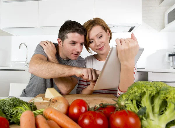 American couple in domestic kitchen wife following recipe in digital pad working together with husband