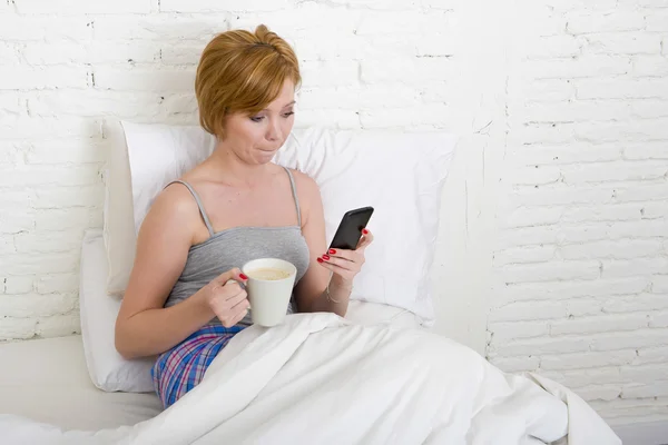 Young beautiful girl having coffee on bed while using internet mobile phone in online social network