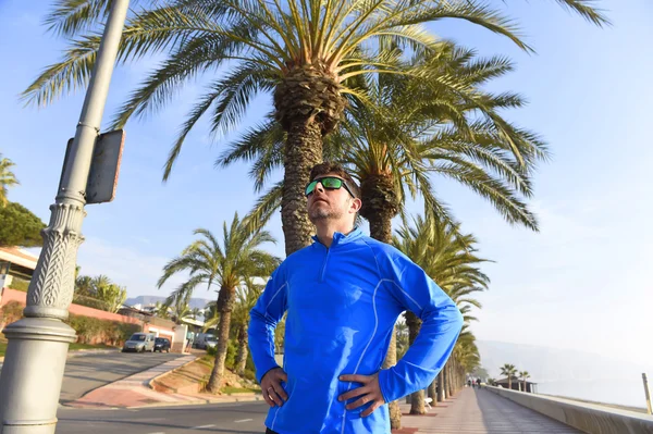 Runner man posing at beach palm trees boulevard with sunglasses in morning jog training session