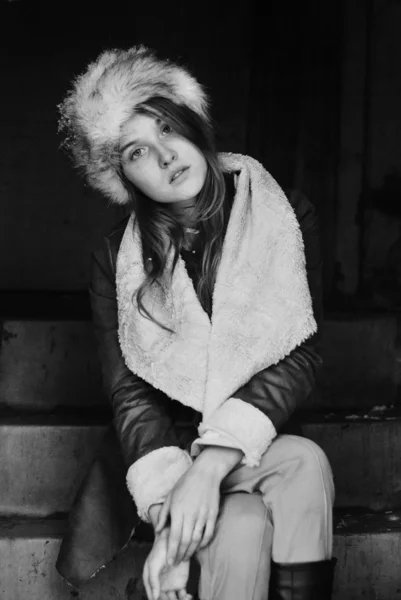 Monochrome portrait photo of posing girl wearing fur hat and leather coat sitting on steps