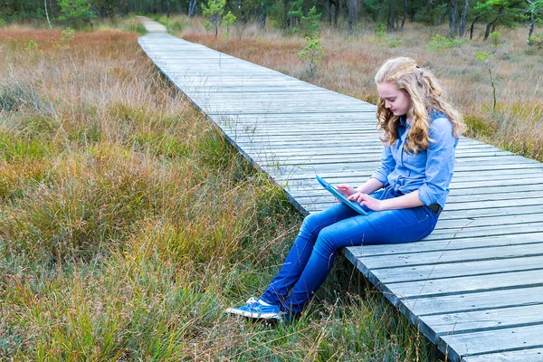 Blonde girl working on tablet computer on wooden path in nature