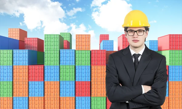 Businessman in yellow helmet with hands crossed, many sea containers of differnt colours behind.