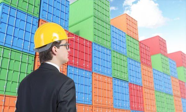 Businessman in yellow helmet looking at many sea containers of differnt colours stocked.