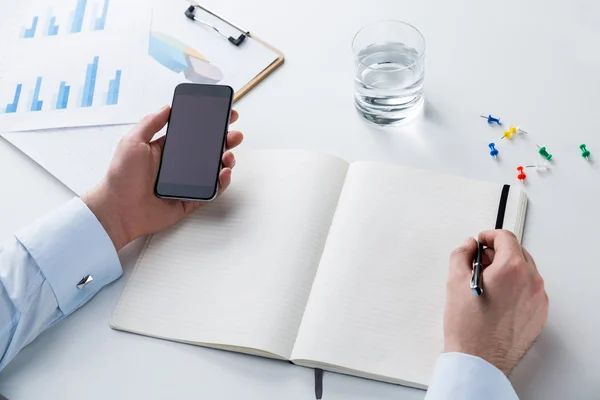 Hands holding smartphone and making notes. Glass of water, pins and graphs on table. Concept of work.