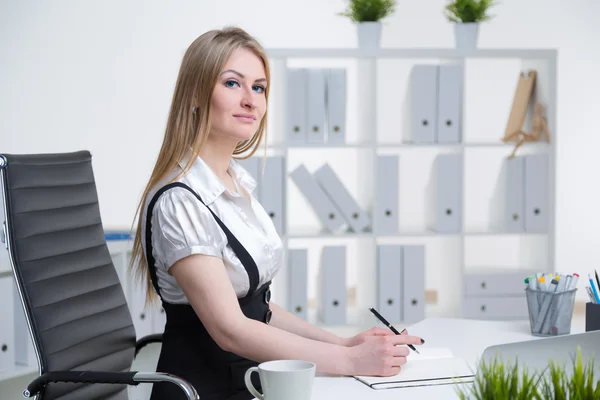 Businesswoman at table making notes. Side view. Shelves at background. Concept of work.