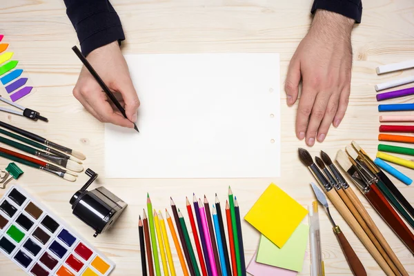 Artist\'s hand drawing on blank white sheet placed on wooden desktop with colorful drawing tools. Mock up