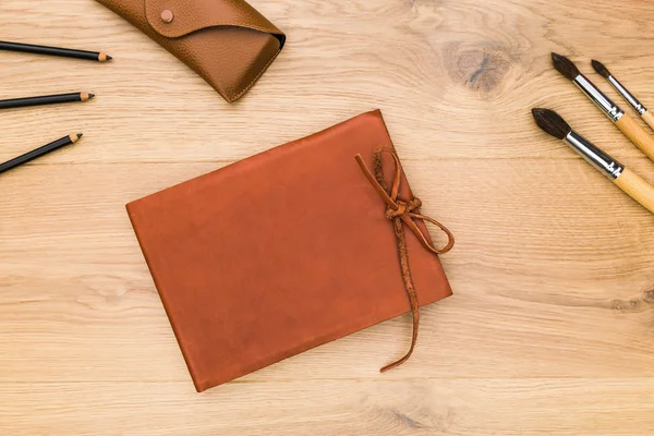 Top view of desktop with closed leather notepad, eyeglass case, pencils and paintbrushes