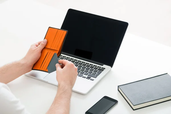 Online shopping concept with businessman taking creadit card out of purse over white desktop with blank laptop screen, book and smart phone. Mock up