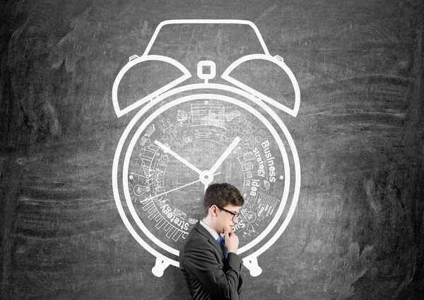 Time management concept with thoughtful businessman against chalkboard wall with alarm clock sketch