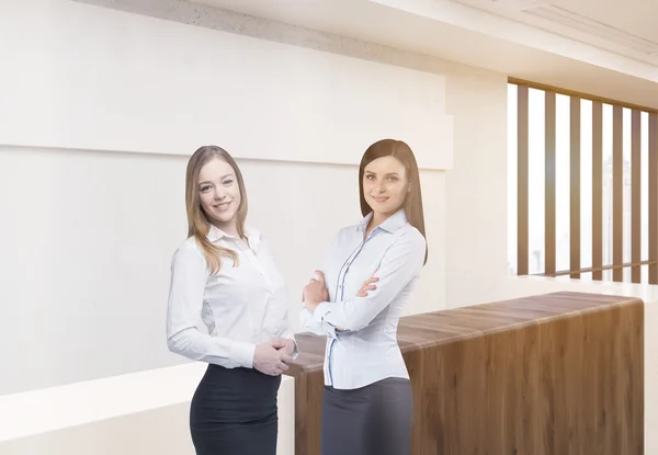 Two attractive european businesswomen standing in front of wooden office reception desk in concrete interior with sunlight. Teamwork concept