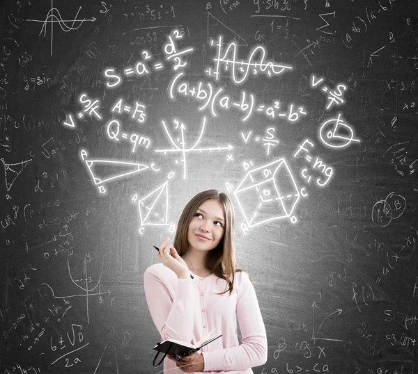 Young woman scientist portrait. Girl with long hair holding notebook and pen looking at formulas above her head and smiling. Concept of woman in engineering