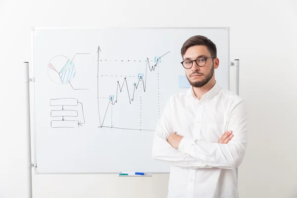 Businessman in glasses with beard is standing near whiteboard with diagrams, his hands crossed. Looking to camera