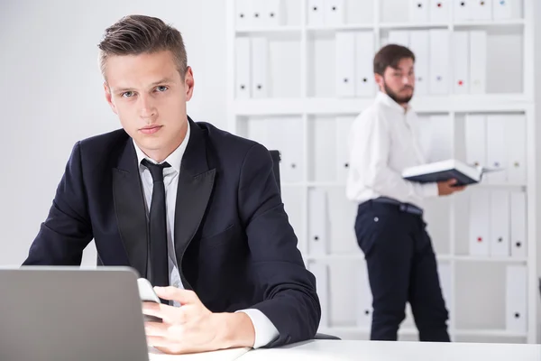 Businessman sitting at his workplace is holding his phone and checking laptop screen. His colleague is standing in background with huge book and looking to camera. Concept of competition in business