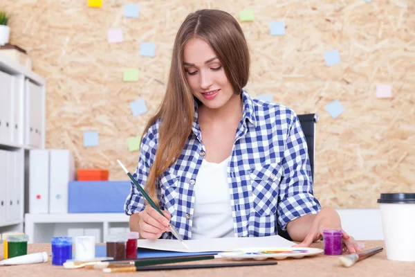 Nice girl with long blond hair is painting in her album. She is sitting at her workplace with table, cork board and book shelves. Concept of sketching for big picture