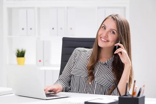 Happy office employee portrait in striped shirt. She is talking on her cell phone at workplace and typing with one hand. Concept of corporate work routine